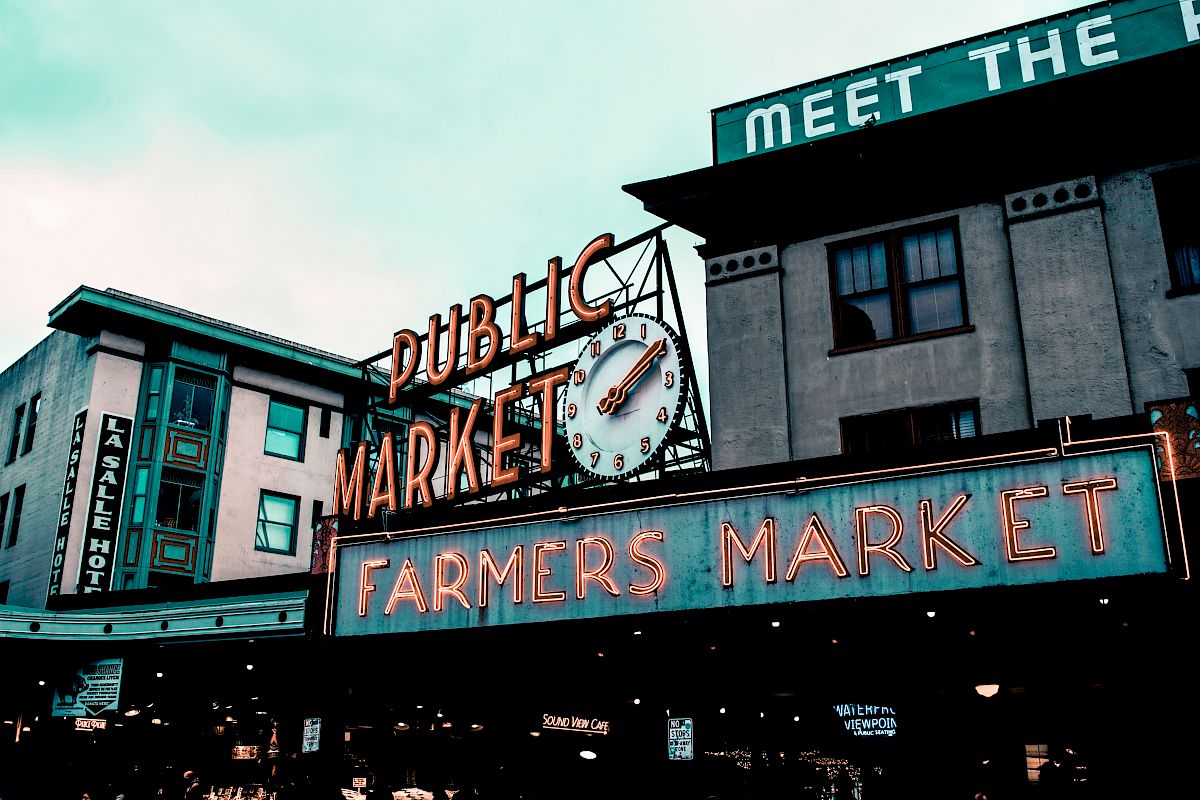 The image shows a public market with a large clock, neon signs reading "Farmers Market," and nearby buildings under a cloudy sky.