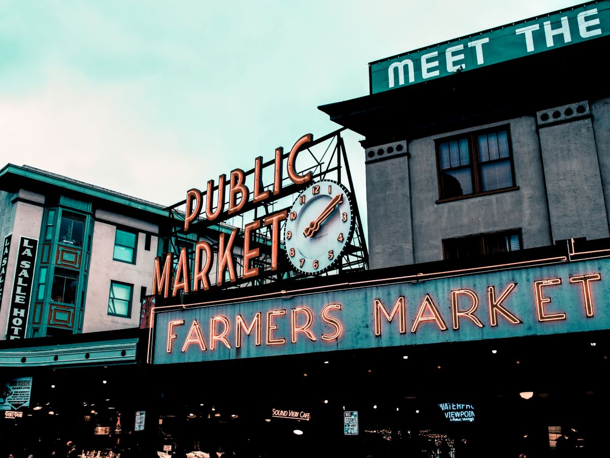 The image shows a public market with a large clock, neon signs reading "Farmers Market," and nearby buildings under a cloudy sky.
