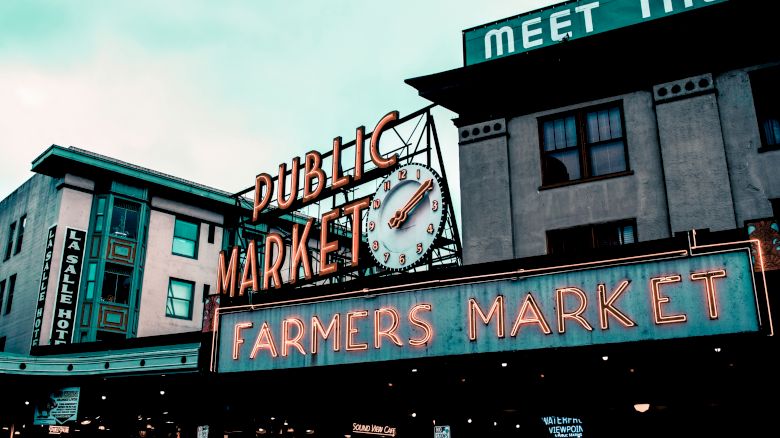 The image shows a public market with a large clock, neon signs reading "Farmers Market," and nearby buildings under a cloudy sky.