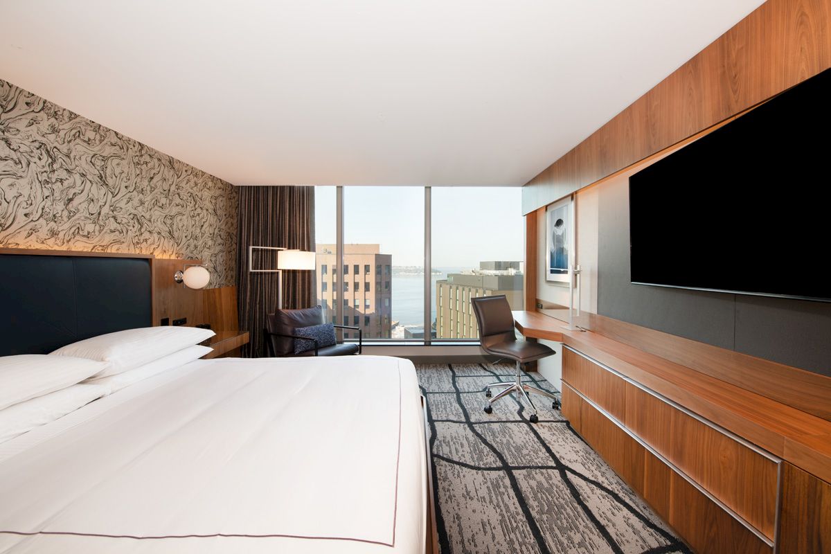A modern hotel room features a large bed, desk, chair, and wall-mounted TV with a window view of distant buildings and the sea.