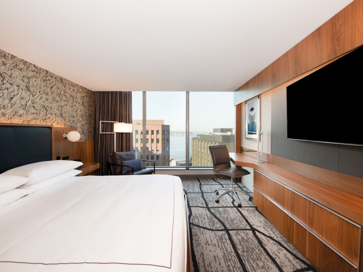 A modern hotel room features a large bed, desk, chair, and wall-mounted TV with a window view of distant buildings and the sea.