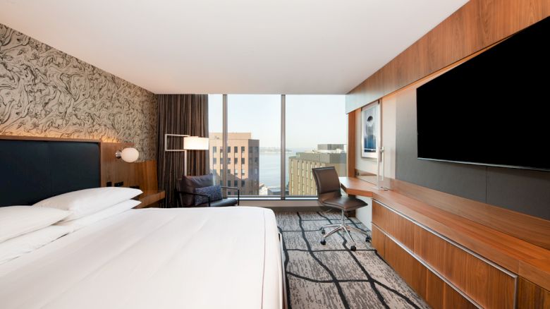 A modern hotel room features a large bed, desk, chair, and wall-mounted TV with a window view of distant buildings and the sea.