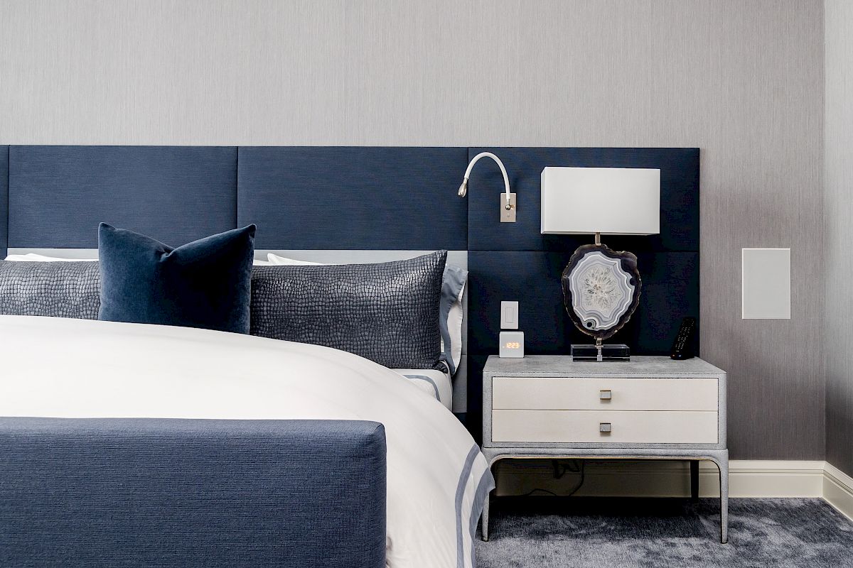 A modern bedroom with a blue headboard, pillows, and a white nightstand featuring a lamp and decorative item beside a neatly made bed.