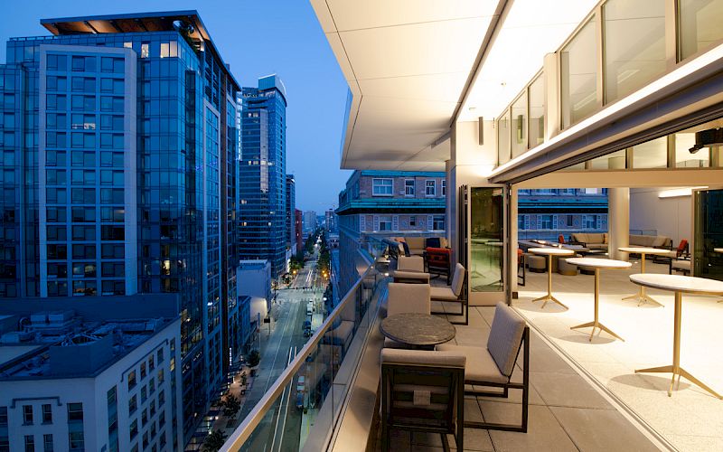 A modern cityscape view from a stylish rooftop terrace at dusk, featuring outdoor seating and tables, overlooking lit-up buildings and street.